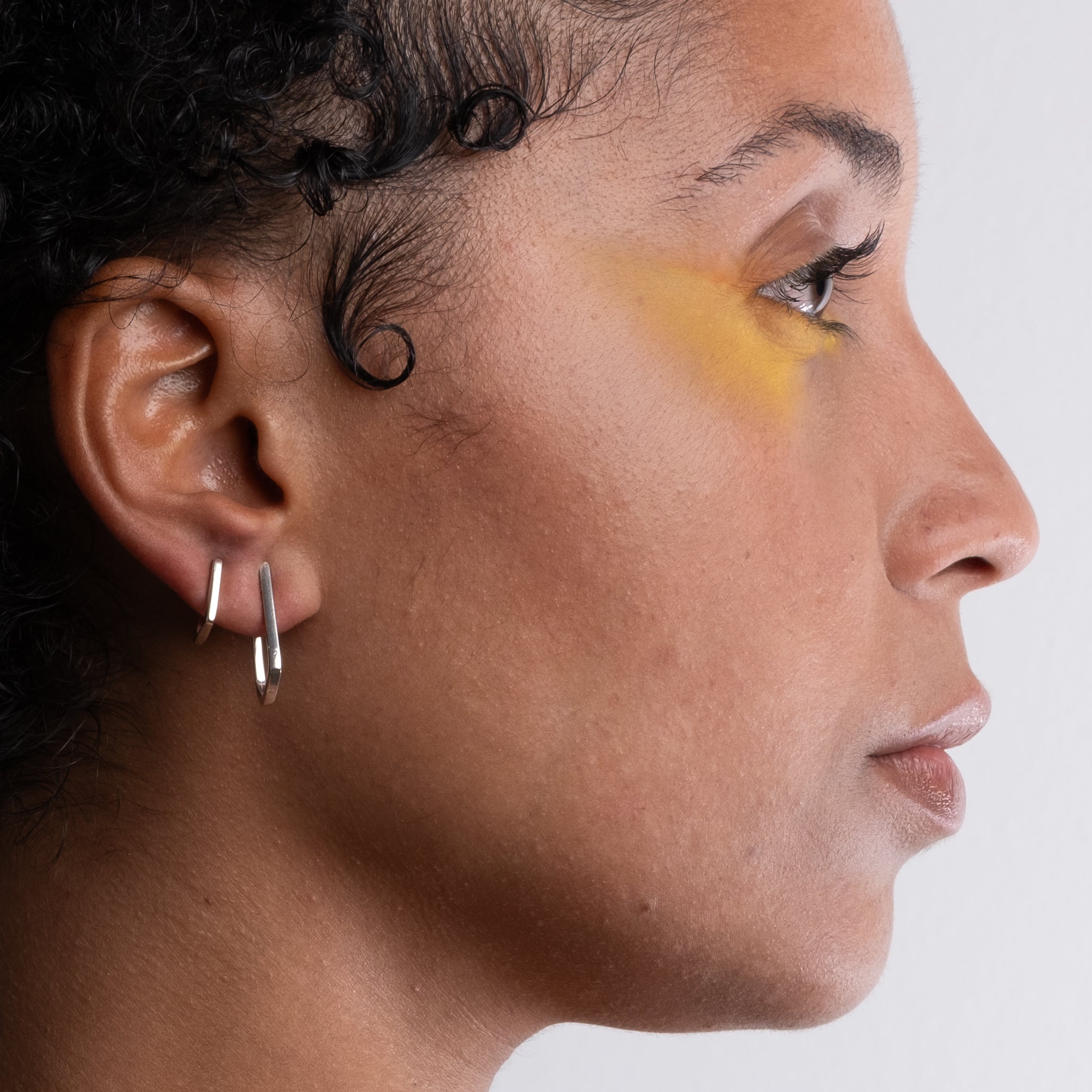 Side profile of a woman with a yellow mark on her face against a white background wearring silver earrings
