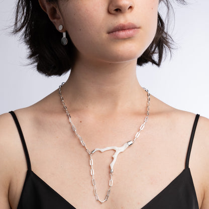A close-up of a model wearing a silver Y-shaped chain necklace with a central white coral, paired with a black spaghetti-strap top against a light gray background.