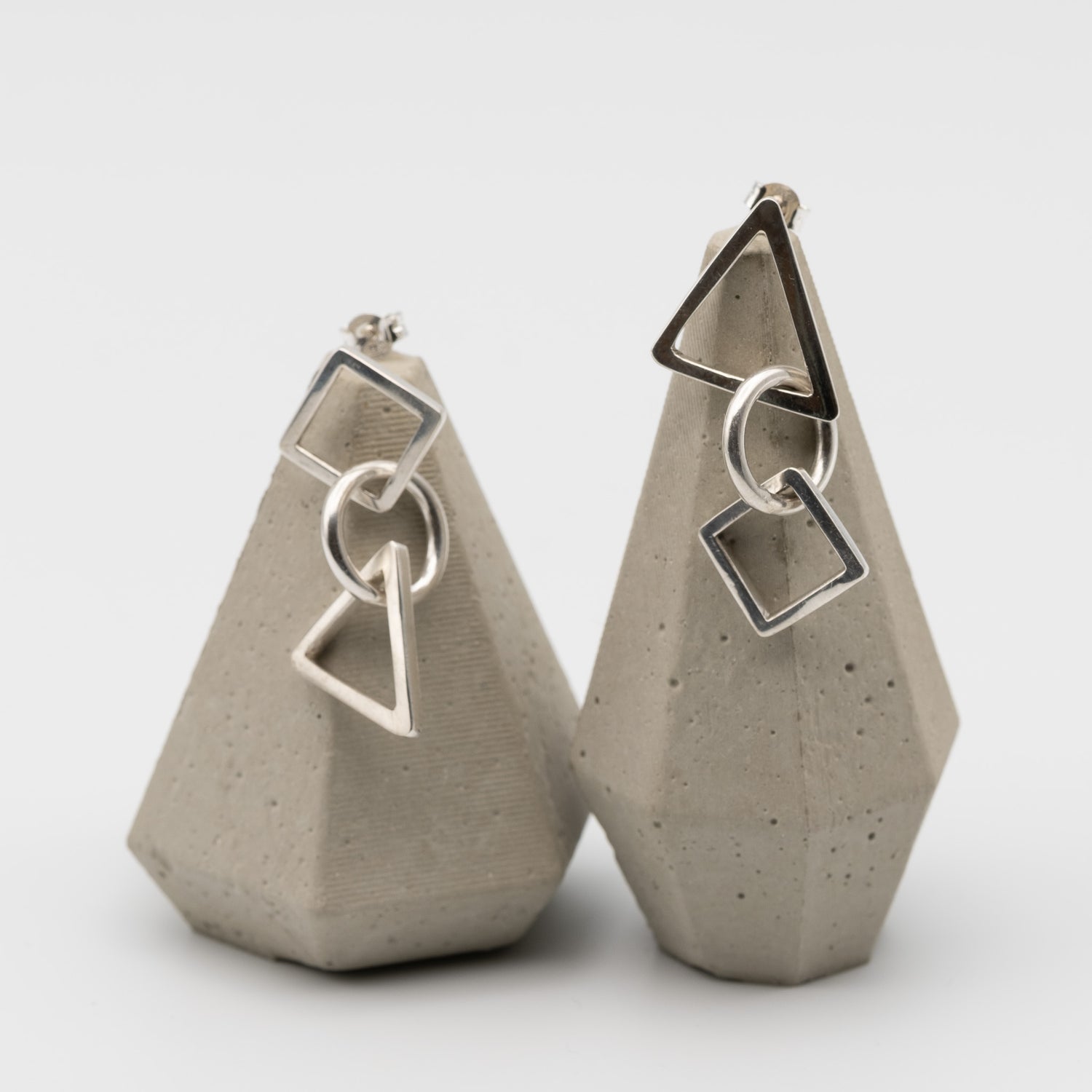A pair of silver geometric dangle earrings displayed on textured gray concrete stands against a clean white background.