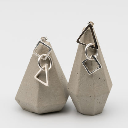 A pair of silver geometric dangle earrings displayed on textured gray concrete stands against a clean white background.