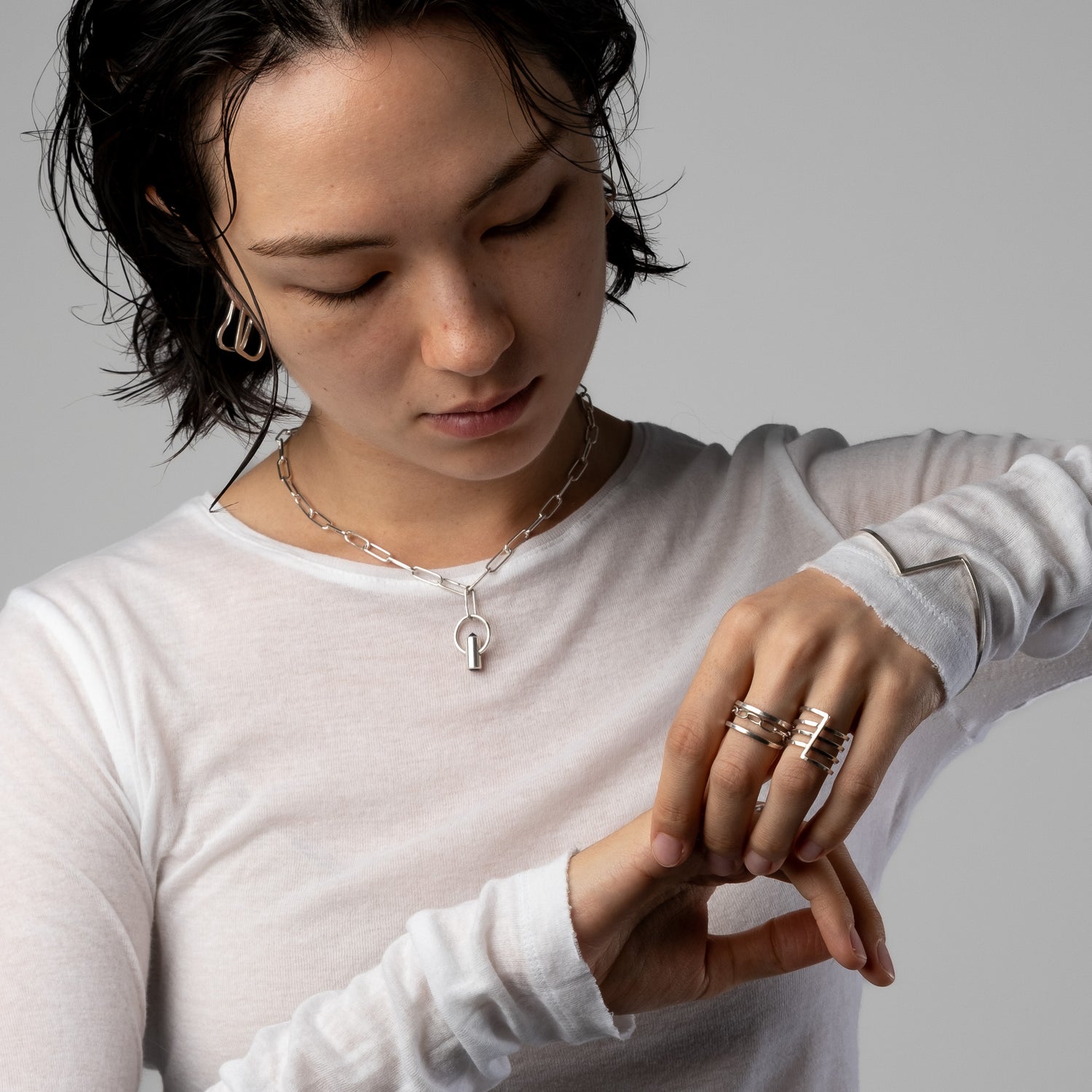 A close-up lifestyle shot of a model wearing a silver key-shaped pendant necklace and several stacked rings on her fingers, wearing a white long-sleeve shirt against a neutral gray background.