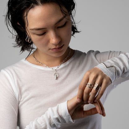 A close-up lifestyle shot of a model wearing a silver key-shaped pendant necklace and several stacked rings on her fingers, wearing a white long-sleeve shirt against a neutral gray background.