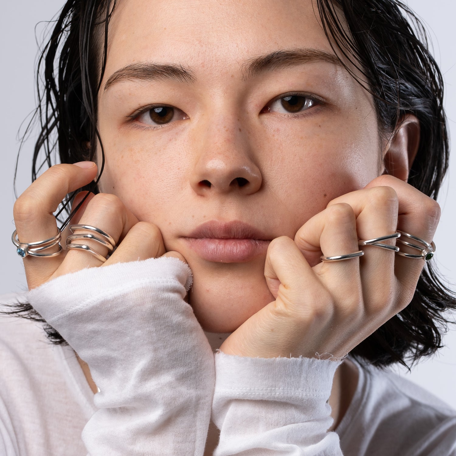 A close-up portrait of a person with wet dark hair resting their chin on their hands, wearing multiple thin silver rings on their fingers and wrists. They are wearing a white long-sleeve shirt against a light gray background.