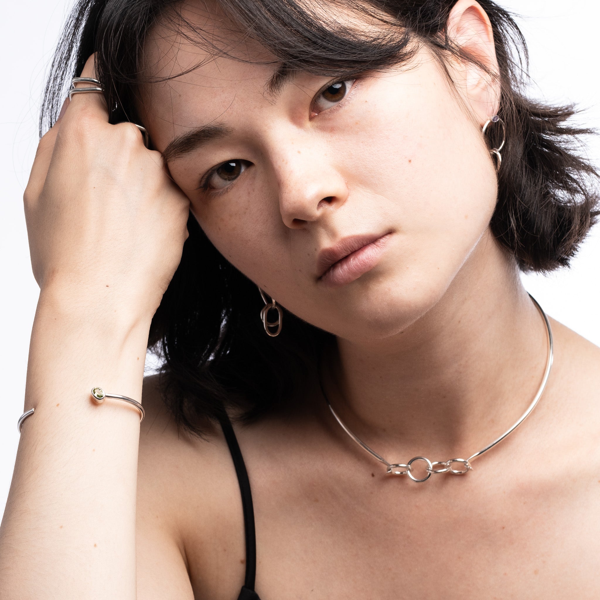 A close-up portrait of a woman wearing delicate silver jewelry, including a thin chain necklace with a double-ring motif, hoop earrings, and a bangle bracelet, against a clean white background.