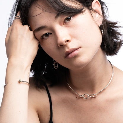 A close-up portrait of a woman wearing delicate silver jewelry, including a thin chain necklace with a double-ring motif, hoop earrings, and a bangle bracelet, against a clean white background.