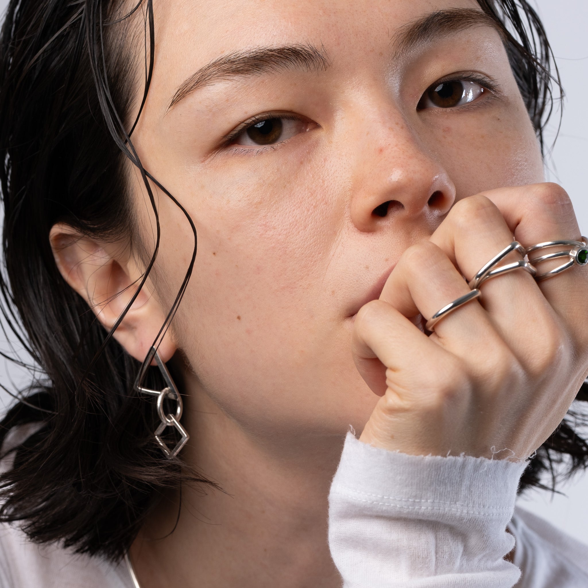 Close-up of a person wearing silver geometric chain-link earrings and matching multi-band rings, with dark hair and a white top against a light gray background.