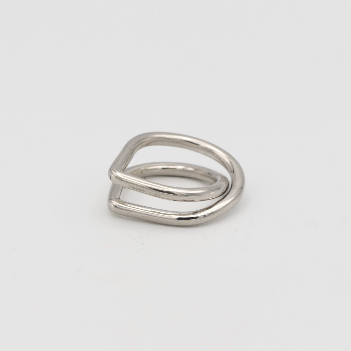 A minimalist silver ring with a unique, open geometric design resembling a double loop or a stylized letter, photographed on a clean light gray background.