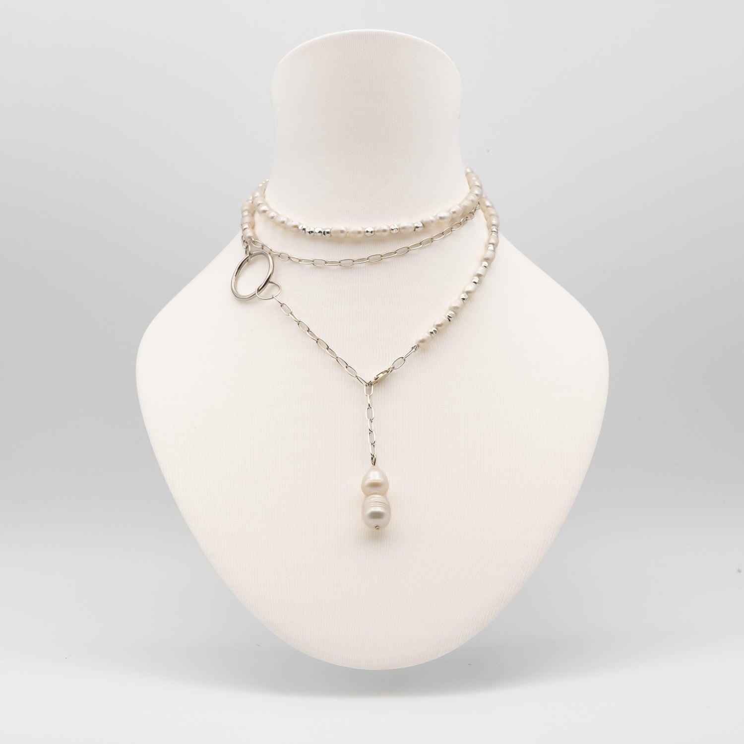 A layered silver-tone necklace displayed on a white mannequin bust. The necklace features a beaded choker layer, a delicate chain layer with a small silver ring accent, and a longer chain with a single white pearl pendant.