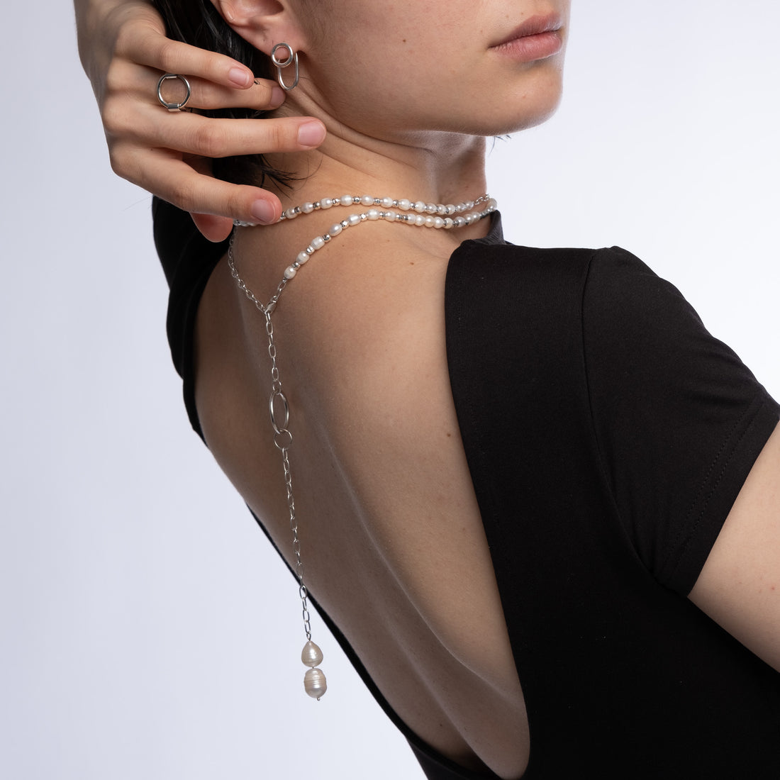 A close-up of a model from the back wearing a delicate silver chain choker adorned with small pearls and a long drop chain with two pearl beads, paired with a black top.