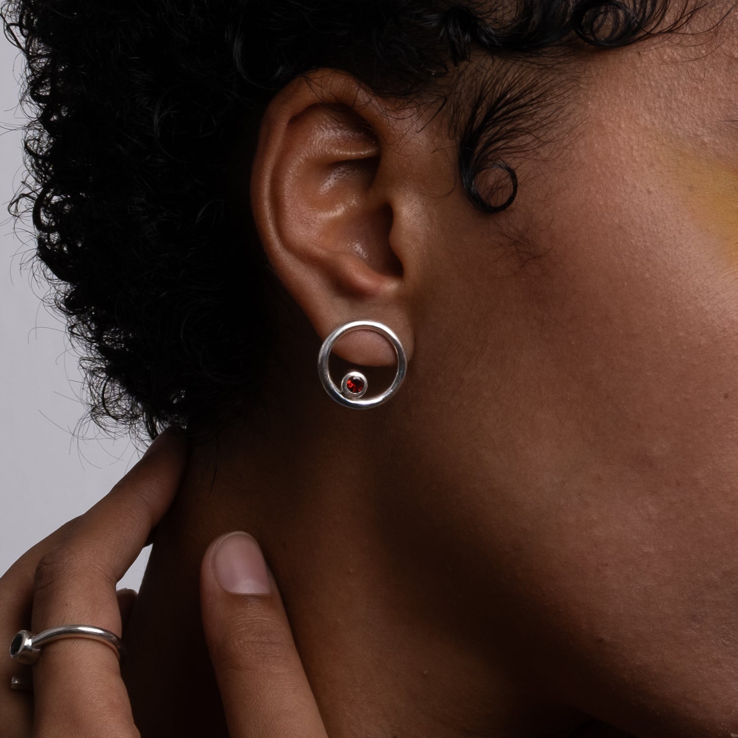 Close-up of an ear wearing a silver earring with a red gemstone, against a neutral background.
