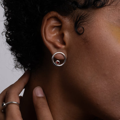 Close-up of an ear wearing a silver earring with a red gemstone, against a neutral background.