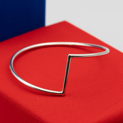 A polished silver  bracelet with a sharp geometric V-shaped corner, displayed on a textured red surface.