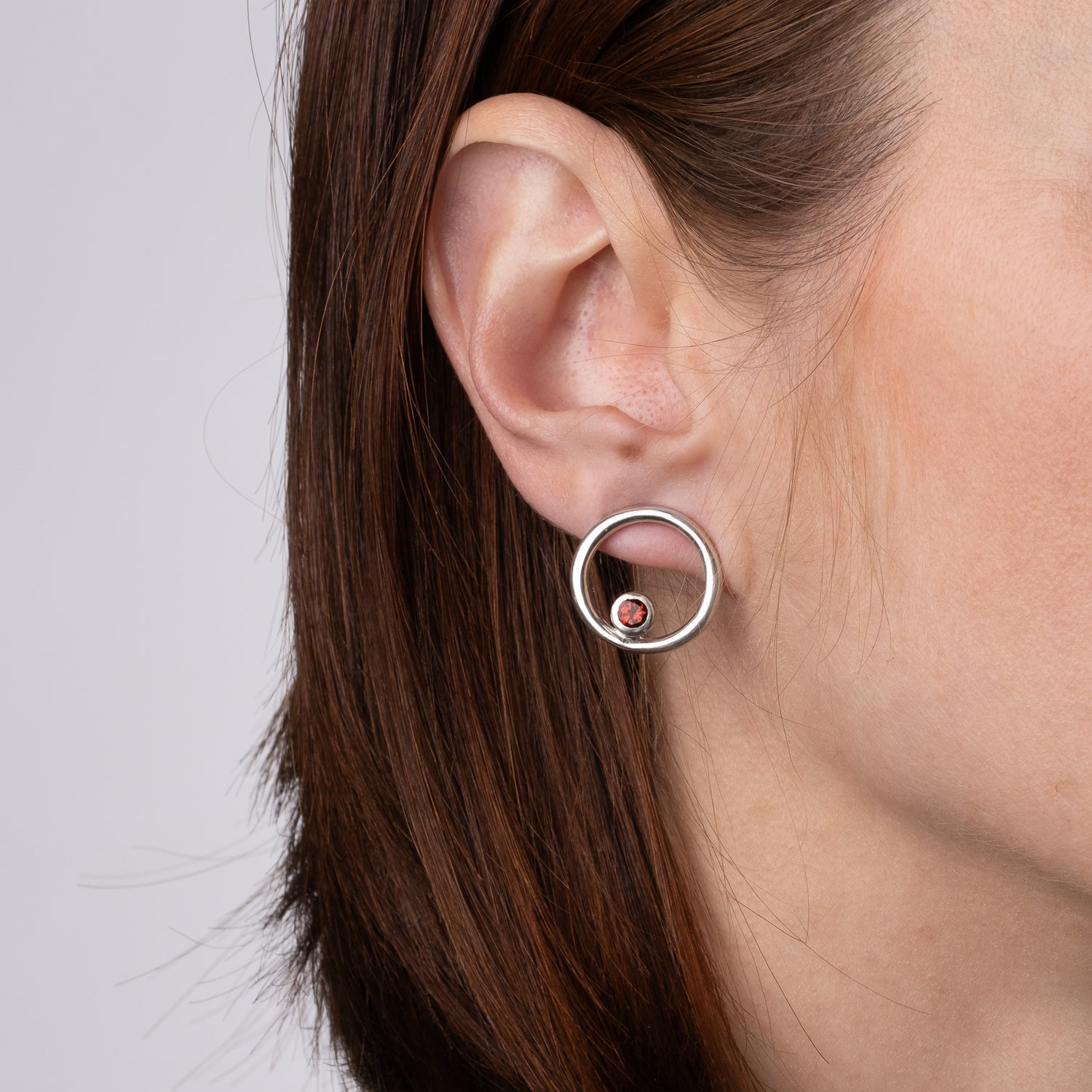Close-up of an ear wearing a silver earring with a red gemstone on a neutral background