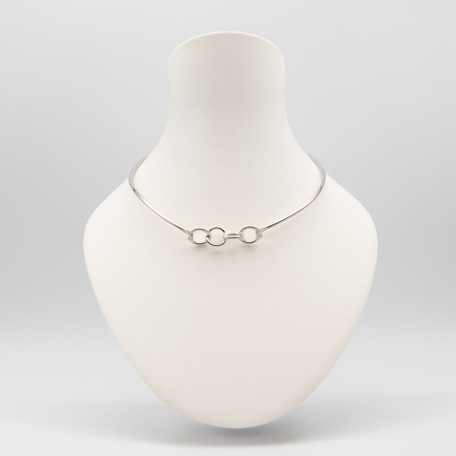 A delicate silver chain necklace with three interlocking circular links, displayed on a matte white jewelry bust against a light gray background.