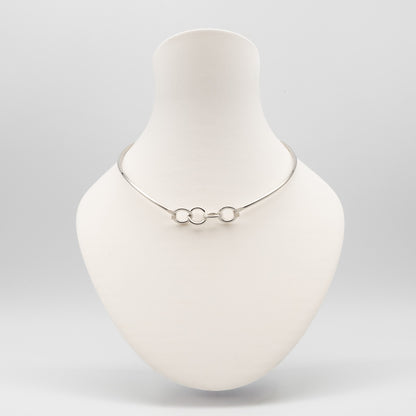 A delicate silver chain necklace with three interlocking circular links, displayed on a matte white jewelry bust against a light gray background.