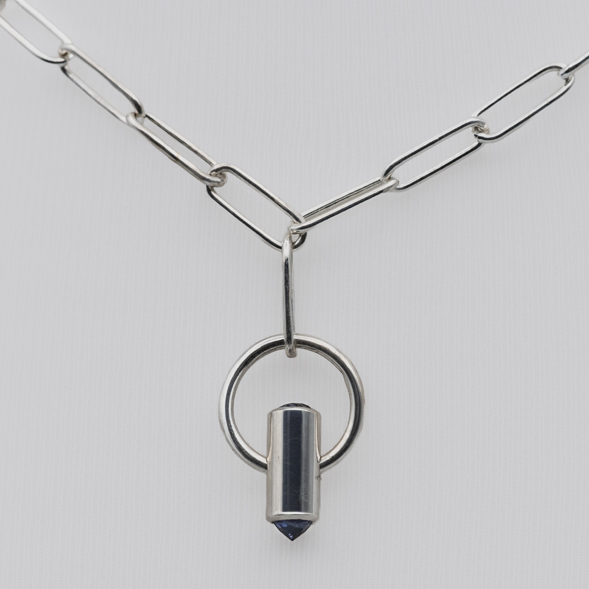 A silver paperclip chain necklace featuring a pendant with a circular ring and a polished cylindrical bullet-shaped charm with a dark blue faceted tip, set against a light gray background.