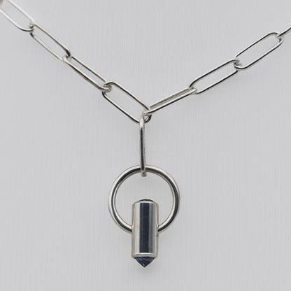A silver paperclip chain necklace featuring a pendant with a circular ring and a polished cylindrical bullet-shaped charm with a dark blue faceted tip, set against a light gray background.