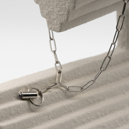 A close-up of a silver chain necklace draped over a textured light gray surface, with a bullet-shaped pendant and ring clasp visible.