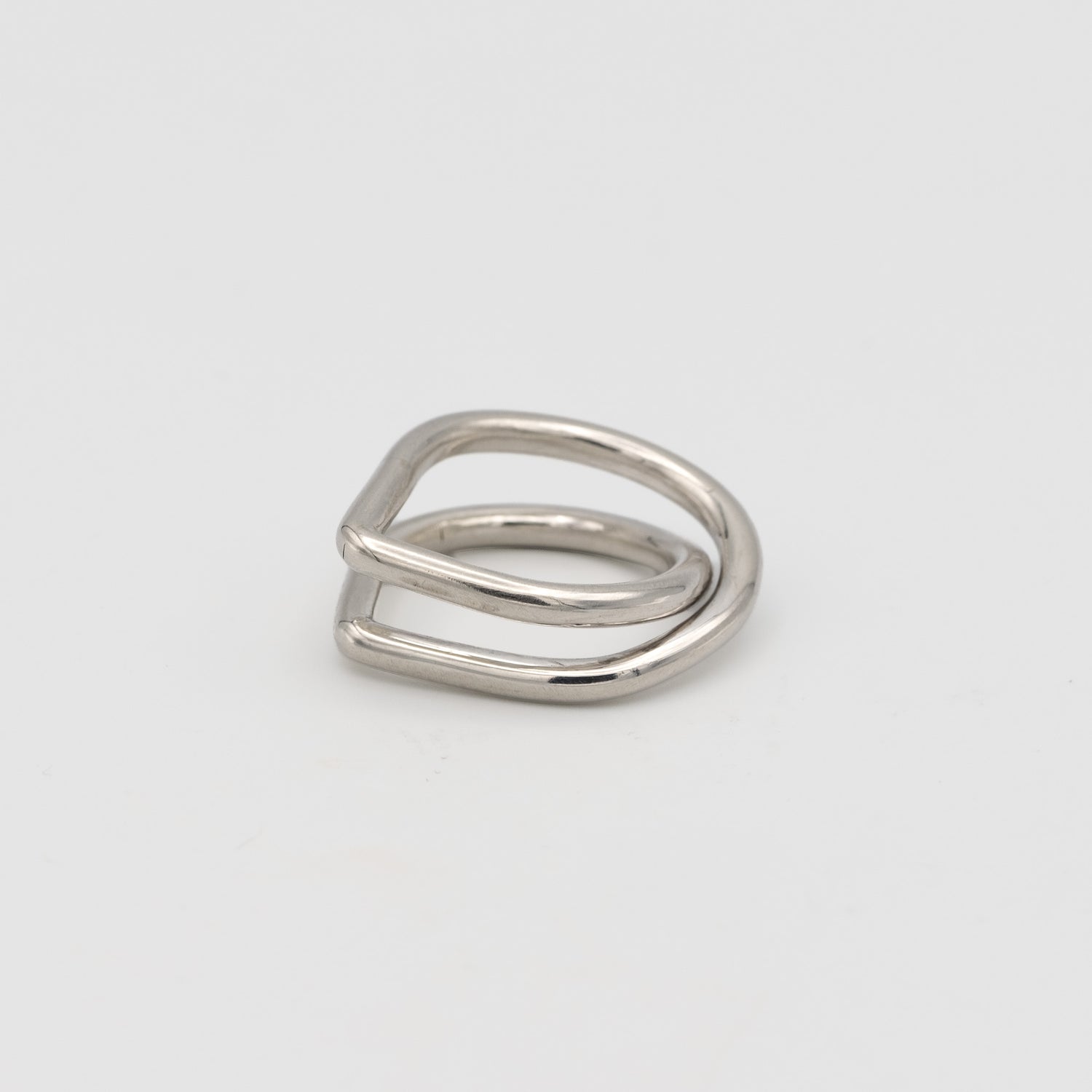 A minimalist silver ring with a unique, open geometric design resembling a double loop or a stylized letter, photographed on a clean light gray background.