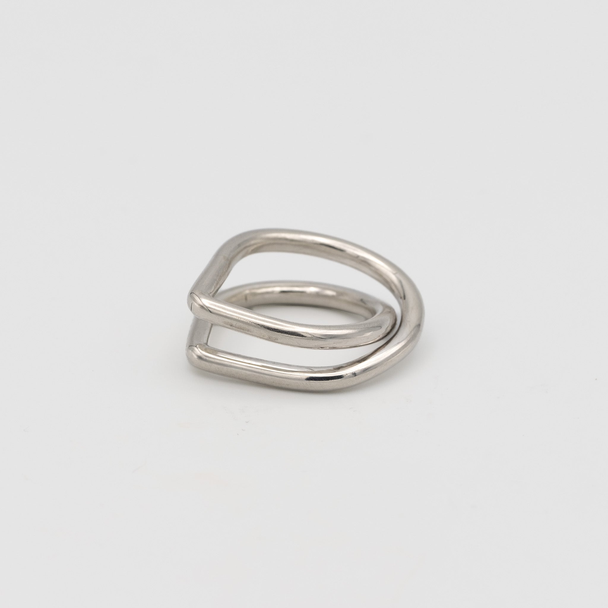 A minimalist silver ring with a unique, open geometric design resembling a double loop or a stylized letter, photographed on a clean light gray background.