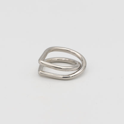 A minimalist silver ring with a unique, open geometric design resembling a double loop or a stylized letter, photographed on a clean light gray background.